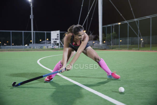 Determined young female field hockey player hitting the ball on field at night — Stock Photo