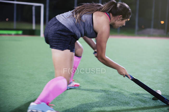 Determined young female field hockey player hitting the ball, playing on field at night — Stock Photo