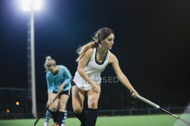 Focused young female field hockey player reaching with hockey stick, practicing on field at night — Stock Photo