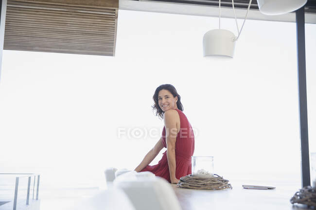 Portrait femme brune souriante et confiante en robe rouge assise à la table à manger — Photo de stock