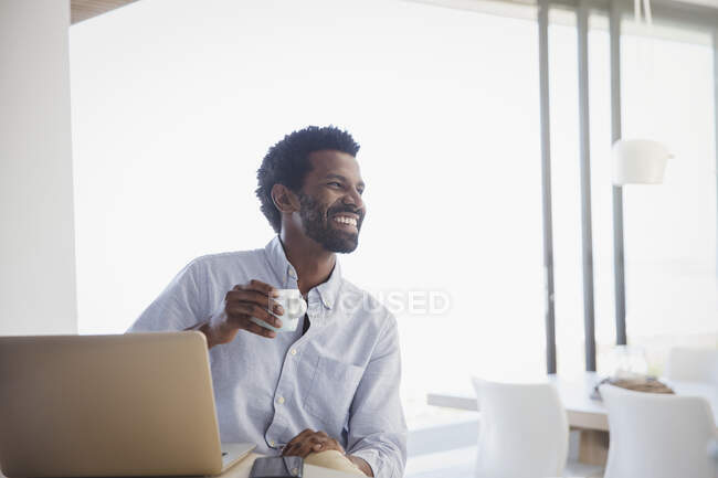 Homme souriant et enthousiaste buvant du café et travaillant à l'ordinateur portable à la maison — Photo de stock