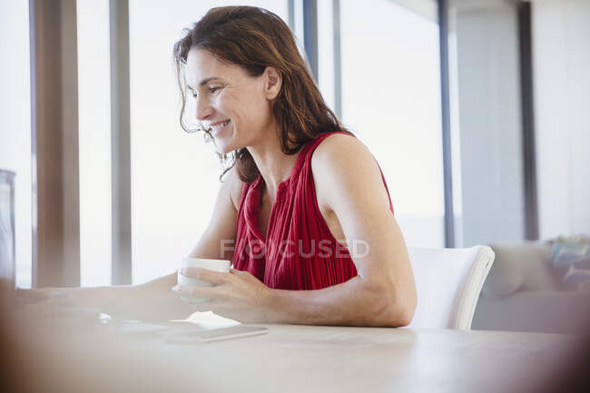 Sonriente morena bebiendo café y trabajando en la mesa de comedor - foto de stock