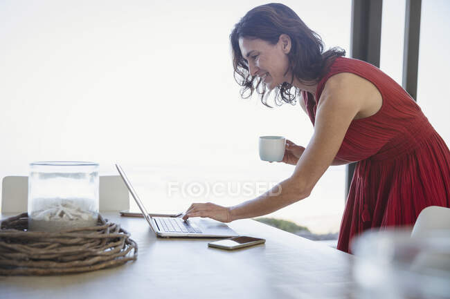 Mujer morena bebiendo café y utilizando el ordenador portátil en la mesa de comedor - foto de stock