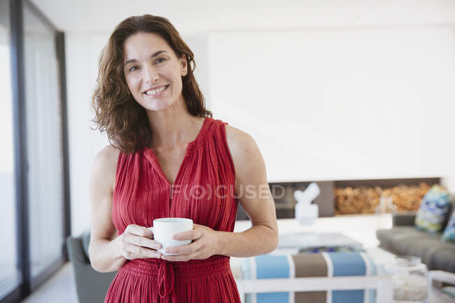 Retrato sonriente, mujer morena confiada bebiendo café en la sala de estar - foto de stock
