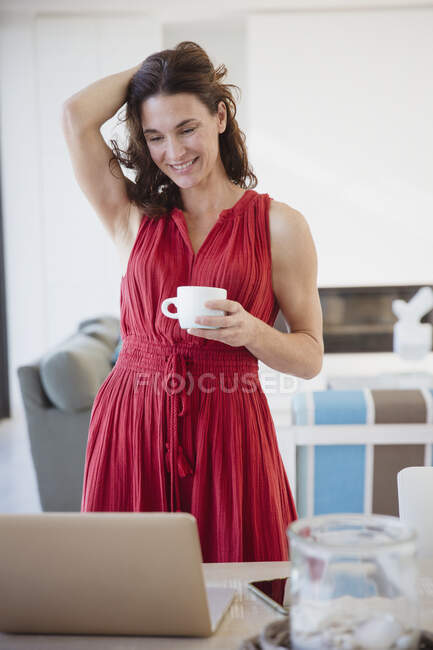 Sorridente donna bruna che beve caffè, lavora al computer portatile in sala da pranzo — Foto stock