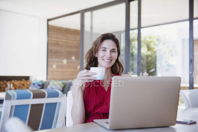 Retrato sonriente, mujer morena confiada bebiendo café y utilizando el ordenador portátil en el comedor - foto de stock