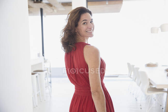 Retrato sonriente, mujer morena confiada en vestido rojo caminando, mirando por encima del hombro en casa - foto de stock