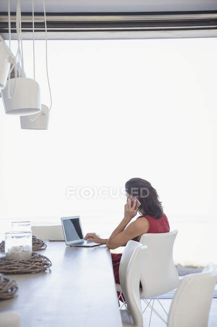 Брюнетка разговаривает по мобильному телефону, использует ноутбук за обеденным столом — стоковое фото