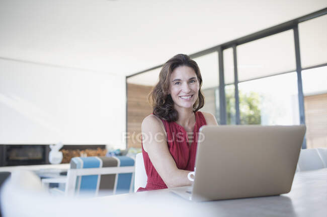 Retrato sonriente morena mujer trabajando en el portátil en la cocina - foto de stock