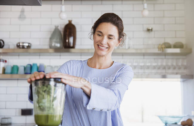 Mulher sorrindo fazendo smoothie verde saudável no liquidificador na cozinha — Fotografia de Stock