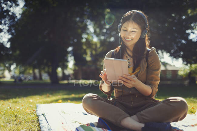 Mujer joven sonriente usando tableta digital, escuchando música con auriculares en el soleado parque de verano - foto de stock