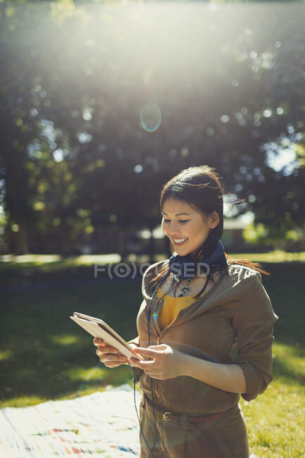 Усміхнена молода жінка з навушниками, використовуючи цифровий планшет у сонячному літньому парку — стокове фото