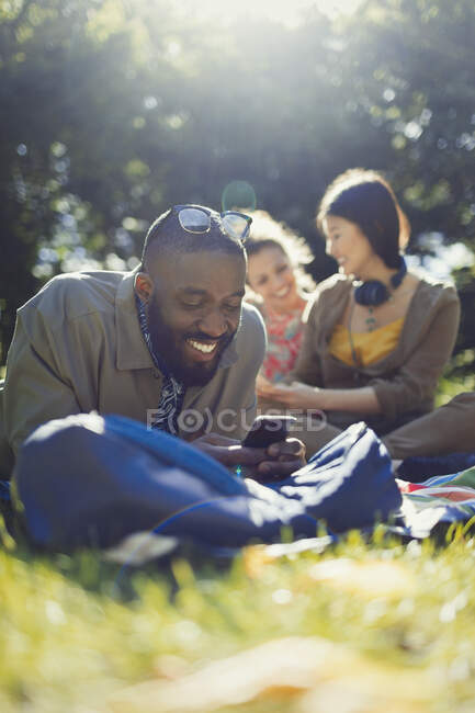 Sonriendo joven hombre mensajes de texto con el teléfono celular y relajarse en el soleado parque de verano - foto de stock