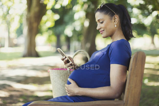 Mujer embarazada mensajes de texto con teléfono celular en el banco del parque - foto de stock