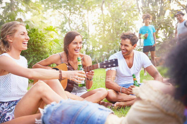 Jóvenes amigos riendo, pasando el rato bebiendo y tocando la guitarra en el parque de verano - foto de stock