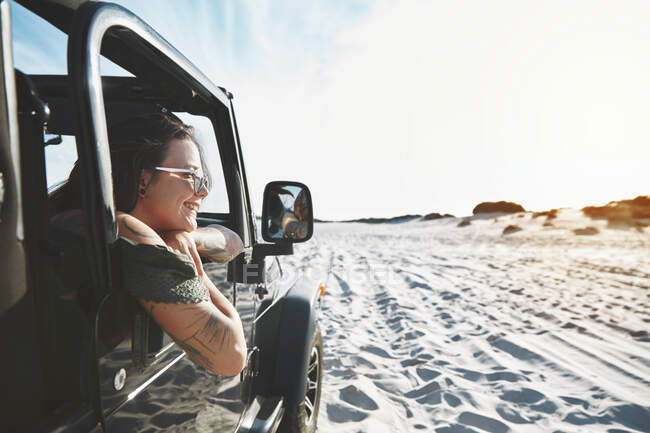 Smiling young woman leaning out jeep window, enjoying beach road trip — Stock Photo