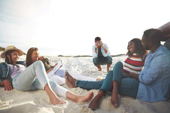 Jovem com câmera digital fotografando amigos relaxando na praia — Fotografia de Stock
