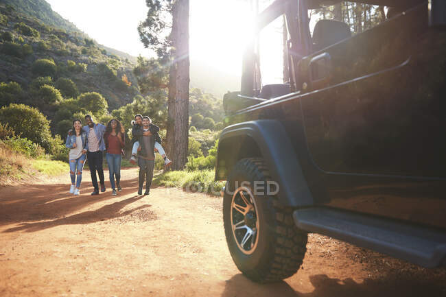 Young friends walking toward jeep on sunny dirt road in woods — Stock Photo