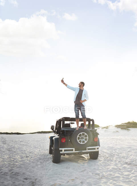 Giovane uomo con fotocamera telefono scattare selfie sulla parte superiore della jeep pneumatico sulla spiaggia — Foto stock