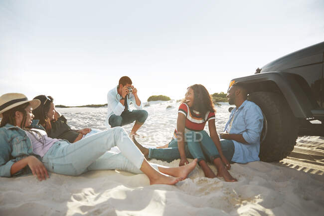 Молодий чоловік з цифровою камерою фотографує друзів, які висять на сонячному пляжі — стокове фото
