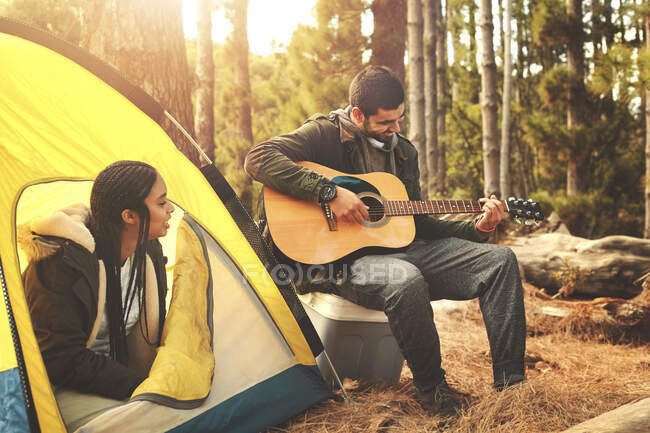 Young couple playing guitar at tent at campsite in woods — Stock Photo