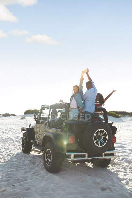 Exuberant young friends cheering with arms raised in jeep on beach, enjoying road trip — Stock Photo
