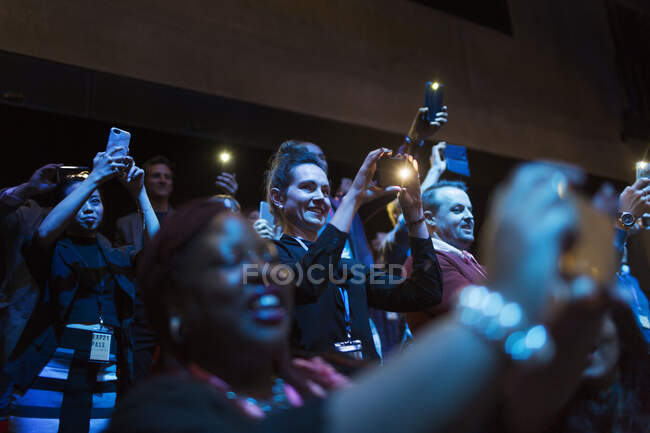 Sonriente y entusiasta audiencia usando teléfonos con cámara en el auditorio oscuro - foto de stock