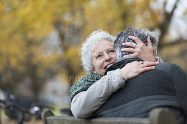 Улыбающаяся, ласковая пожилая пара, обнимающаяся на скамейке запасных в осеннем парке — стоковое фото