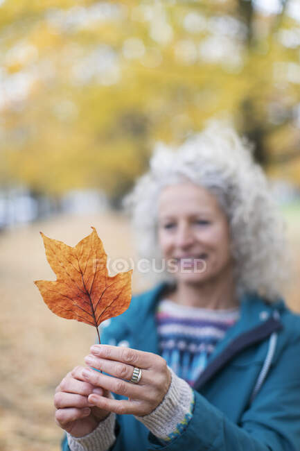 Mulher sênior segurando folha de outono laranja no parque — Fotografia de Stock