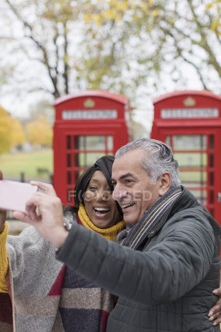 Feliz pareja de ancianos tomando selfie frente a cabinas telefónicas rojas en el parque de otoño - foto de stock