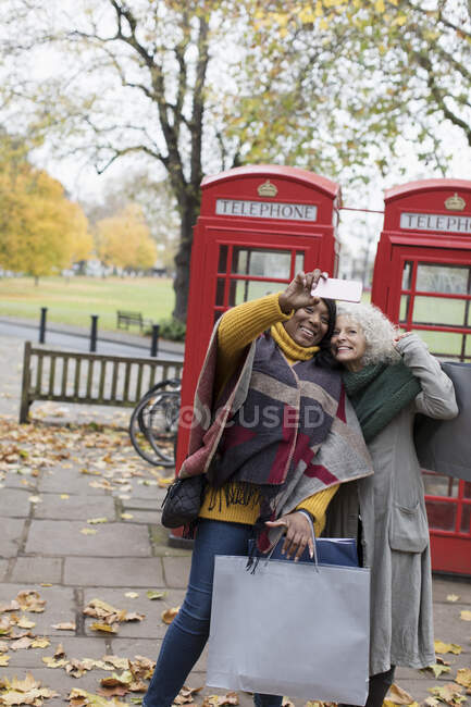 Senior women friends with shopping bags taking selfie in autumn park in front of red telephone booths — Stock Photo
