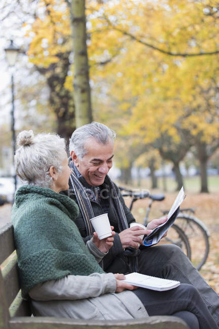 Старша пара читає газету і п'є каву на лавці в осінньому парку — стокове фото