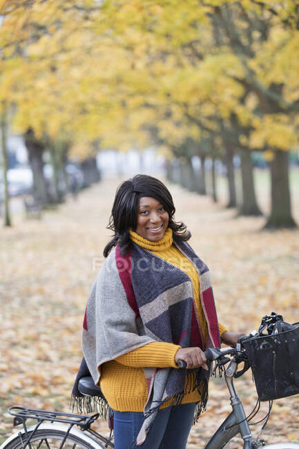Портрет улыбающейся женщины с велосипедом среди листьев и деревьев в осеннем парке — стоковое фото