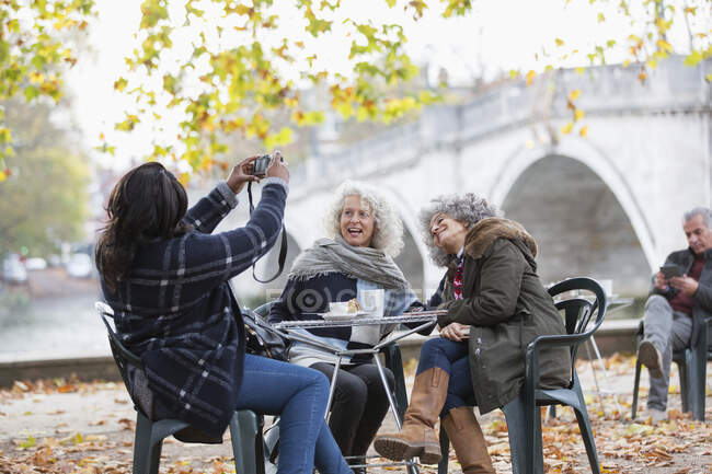 Woman with digital camera photographing active senior women friends at autumn park cafe — Stock Photo