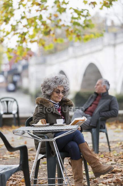 Active senior woman reading book, enjoying cake and coffee at autumn park cafe — Stock Photo
