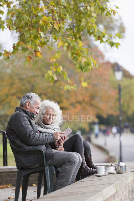 Старша пара використовує цифровий планшет, насолоджуючись кавою і чаєм в осінньому парку — стокове фото