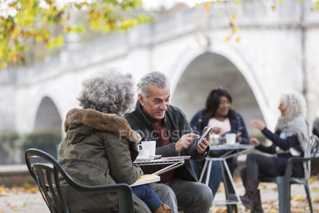 Active senior couple using smart phone, drinking coffee at autumn park cafe — Stock Photo