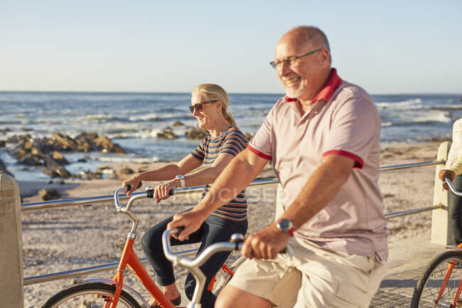 Sonriendo activos turistas senior montar en bicicleta a lo largo del océano soleado - foto de stock