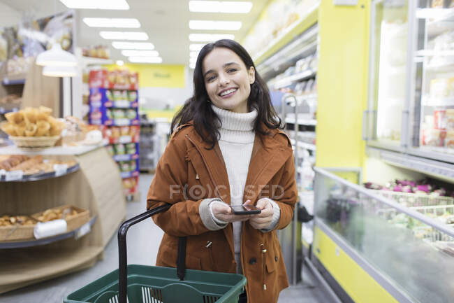 Retrato sorridente, mulher confiante com compras de telefone inteligente no supermercado — Fotografia de Stock
