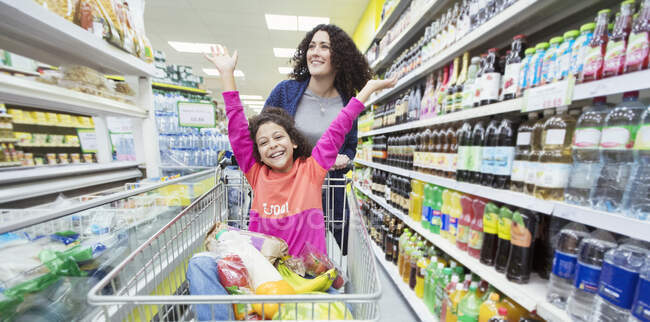 Madre empujando a su hija juguetona en el carrito de compras en el pasillo del supermercado - foto de stock