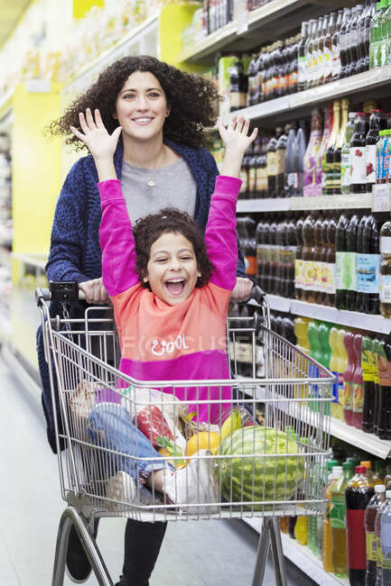 Mother pushing excited daughter in shopping cart in supermarket aisle — Stock Photo
