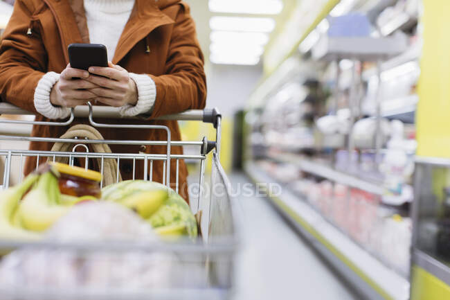 Femme avec téléphone intelligent poussant panier dans le supermarché — Photo de stock