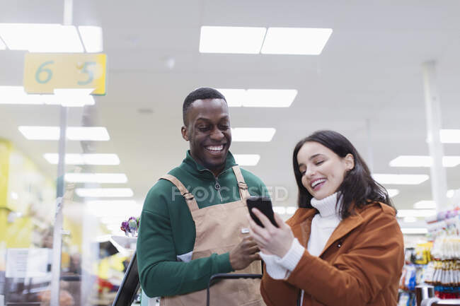 Бакалейщик помогает клиенту со смартфоном в супермаркете — стоковое фото