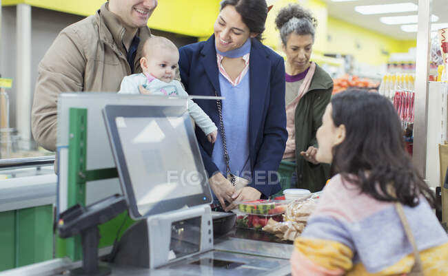 Caixa amigável conversando e ajudando o casal com o bebê no checkout do supermercado — Fotografia de Stock
