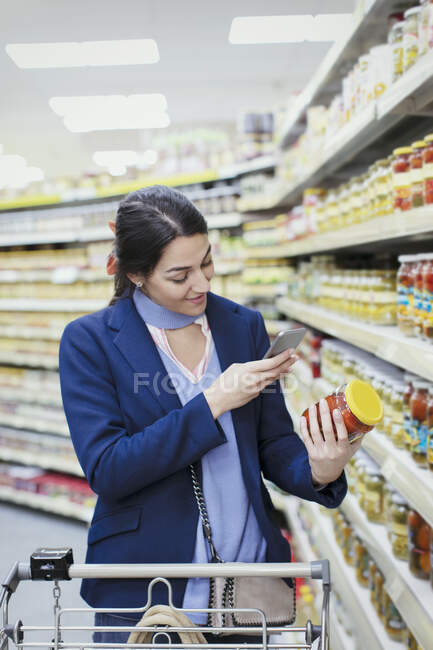 Woman with smart phone scanning label on jar in supermarket — Stock Photo
