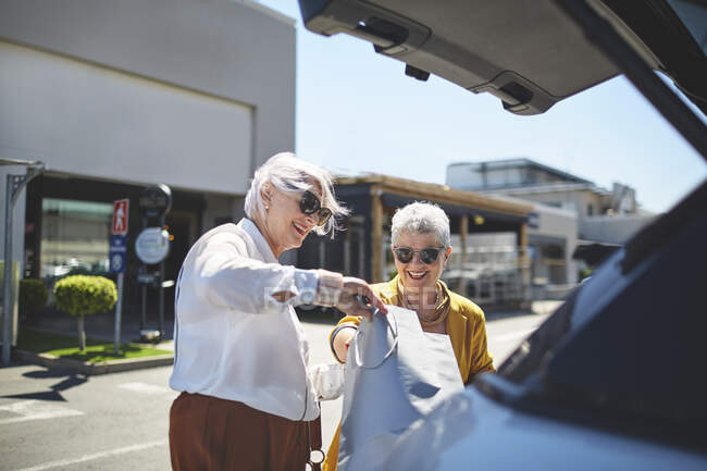 Mujeres mayores amigos cargando bolsas de compras en la parte posterior del coche en el estacionamiento soleado - foto de stock