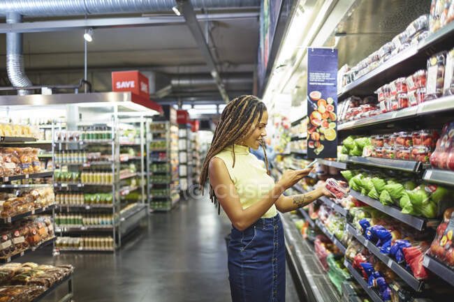 Mujer con compras de teléfonos inteligentes en la sección de productos de supermercado - foto de stock