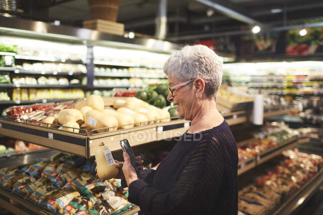Mulher sênior com telefone inteligente de compras em supermercado produzir seção — Fotografia de Stock