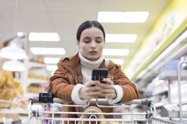 Mujer joven con compras de teléfonos inteligentes en la tienda de comestibles - foto de stock