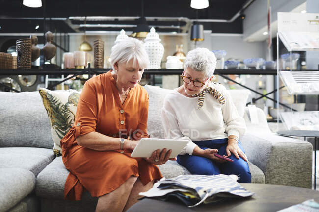 Старшие женщины с цифровой планшет глядя на ткани образцы на диване в мебельном магазине — стоковое фото